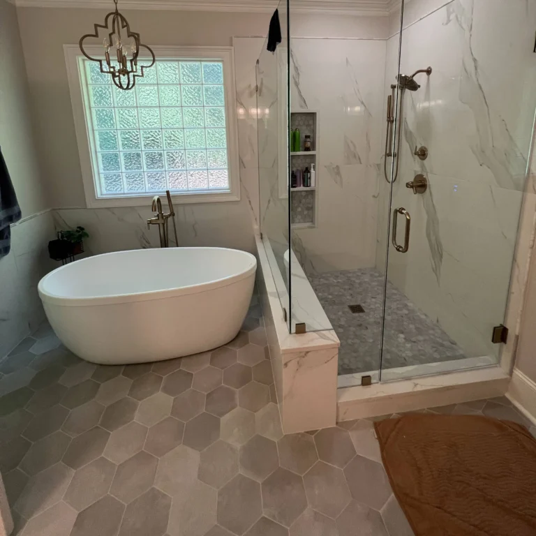Modern bathroom renovation featuring a white freestanding soaking tub beneath a window with turquoise shutters, adjacent to a frameless glass-enclosed walk-in shower with marble-look tile walls and gray tile flooring throughout